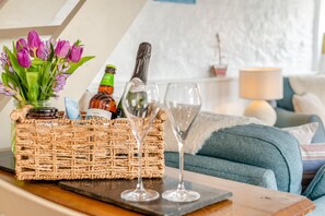 Un panier en osier contenant une bouteille et un verre, posé sur une table, un vase avec des fleurs violettes et, au fond, un canapé bleu.