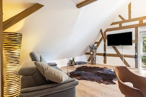 A modern living room with a sloped ceiling, a flat-screen TV mounted on a wooden beam, a leather sofa, a round coffee table, and a rustic rug.
