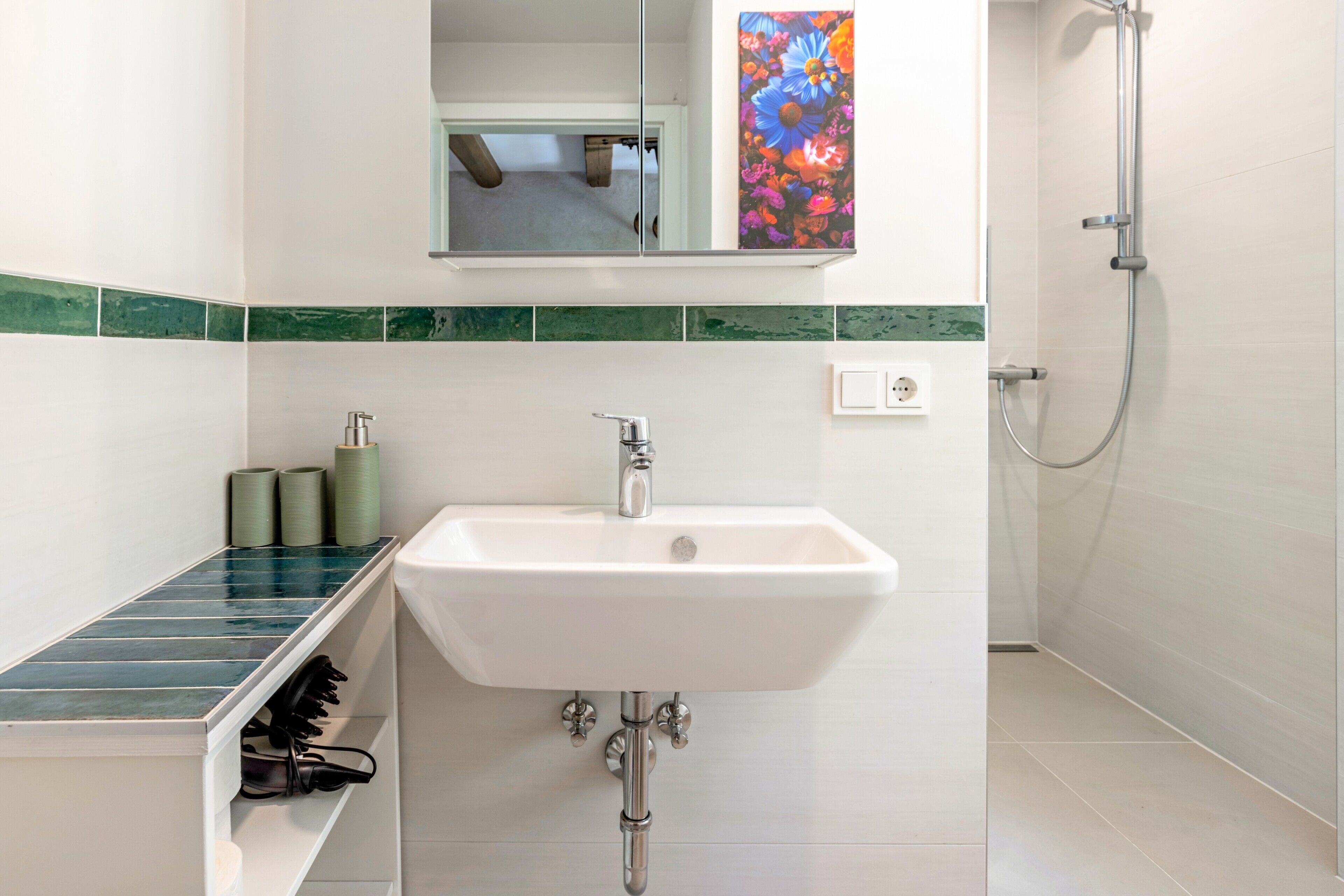 Ein Badezimmer mit einem weißen Waschbecken, einem Spiegel darüber, einer Dusche mit Handbrause und einer grün gefliesten Rückwand.