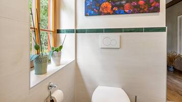 Une salle de bain avec des toilettes, une fenêtre ornée de plantes et un interrupteur fixé au mur.