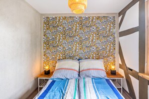 A bedroom with a bed, two bedside tables, a patterned wallpaper, and a hanging light fixture.