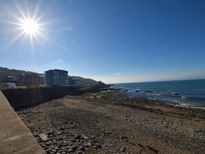 A coastal area with a rocky beach, a concrete barrier, and buildings in the background.