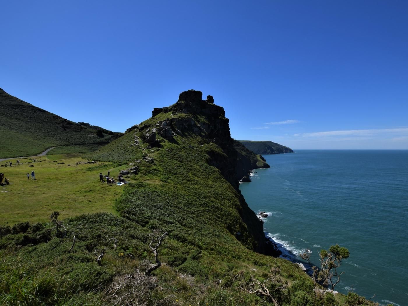 A coastal landscape with a prominent cliff and a grassy area with people.