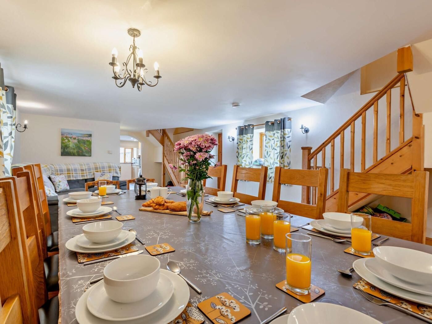 Un coin repas avec une longue table dressée pour le petit-déjeuner, comprenant des assiettes, des bols et des verres de jus d’orange.