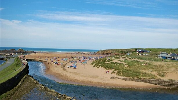 Una playa costera con un río, dunas cubiertas de hierba y algunos edificios al fondo.