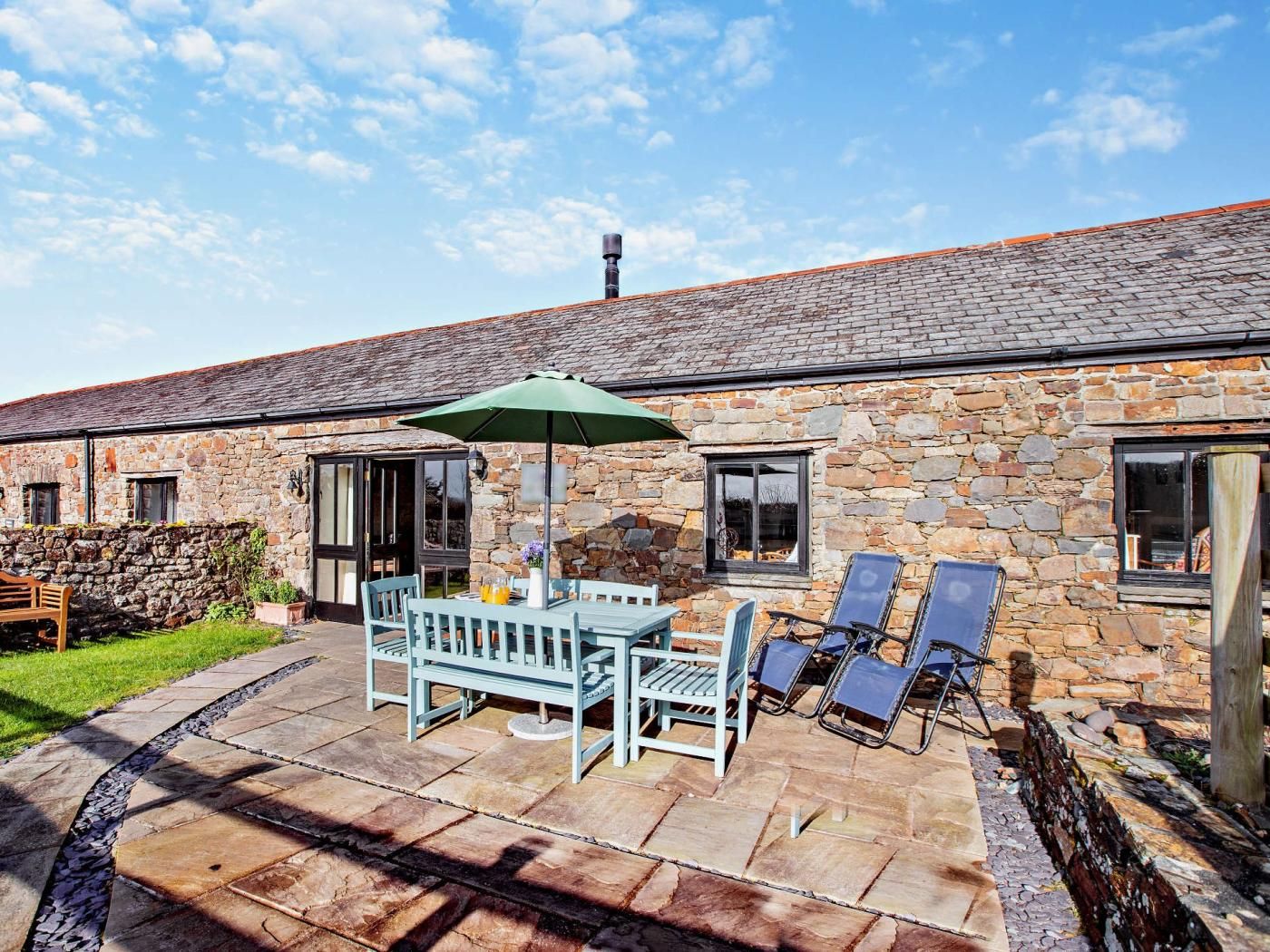 A stone building with a patio area featuring a table and chairs, a green umbrella, and lounge chairs.