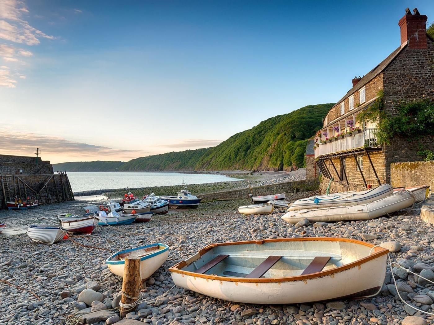 A pebbled beach with several small boats, a stone building with a balcony, and a clear sky.