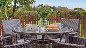 Een ronde rieten tafel met twee witte bekers en een belegd broodje, omringd door rieten stoelen, met een houten hek en groenheid op de achtergrond.