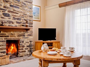 A cozy room with a stone fireplace, a television on a wooden stand, and a round wooden table with plates, cups, and a teapot. - 3 Bed in Plymouth (oc-keepi) (Wembury)