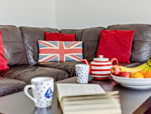Eine braune Ledersofa mit roten Kissen, einem Kissen mit Union Jack Muster, einer weißen Tasse mit blauen Mustern, einem roten und weißen Teekanne und einer Schale mit Obst.