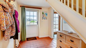 A room with a wooden staircase, a door with a view, a dresser, and a wall-mounted coat rack with jackets. - 3 Bed in Plymouth (oc-combv) (Wembury)