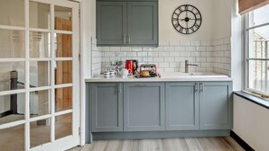A kitchen with grey cabinets, a white countertop, a red kettle, and a wall clock. - 3 Bed in Ilfracombe (oc-haywd) (Ilfracombe)