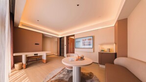 Habitación de hotel moderna con una mesa de comedor redonda, un sofá, un mueble bajo y un televisor montado en la pared.
