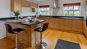 Una cocina moderna con armarios de madera, una isla central con taburetes de bar, una campana extractora de acero inoxidable y ventanas amplias con persianas rojas.