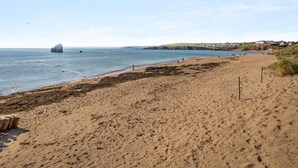 Una playa arenosa con un promontorio rocoso en el agua, cielo despejado y una costa distante con edificios.