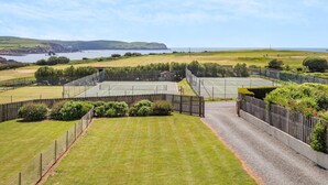 Una cancha de tenis rodeada por una valla, con vista al mar y colinas al fondo.