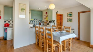 Comedor con una mesa dispuesta para cuatro, sillas de madera, una cocina al fondo y una lámpara en el techo.
