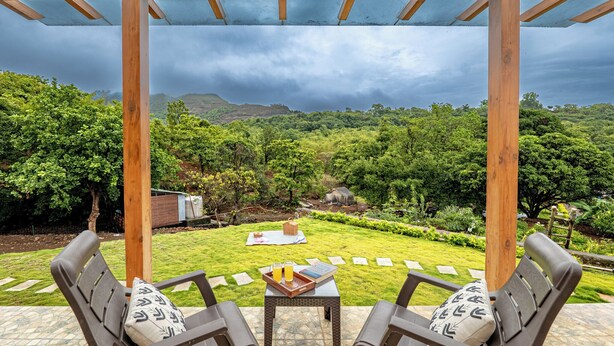 A covered outdoor seating area with a view of a lush garden and distant hills. - CosmicStays Yutori  (Mulshi)