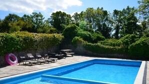 En rektangulär pool med klart, blått vatten, omgiven av en stenbelagd terrass. Längs poolkanten finns solstolar och ett bord med stolar.