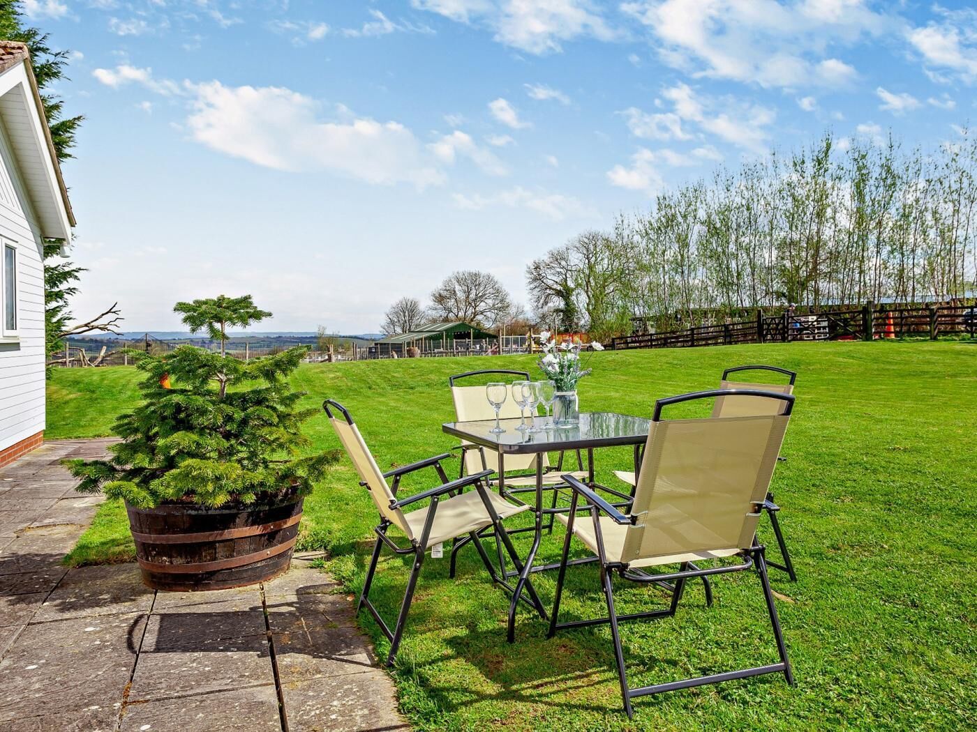 Eine Terrasse mit Tisch und Stühlen, einem Blumentopf und Blick auf ein von Bäumen umgebenes, eingezäuntes Gelände.