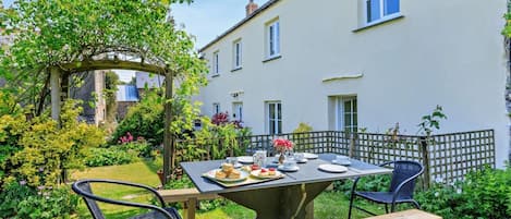 A garden patio with a table set for two, surrounded by greenery and a white building.