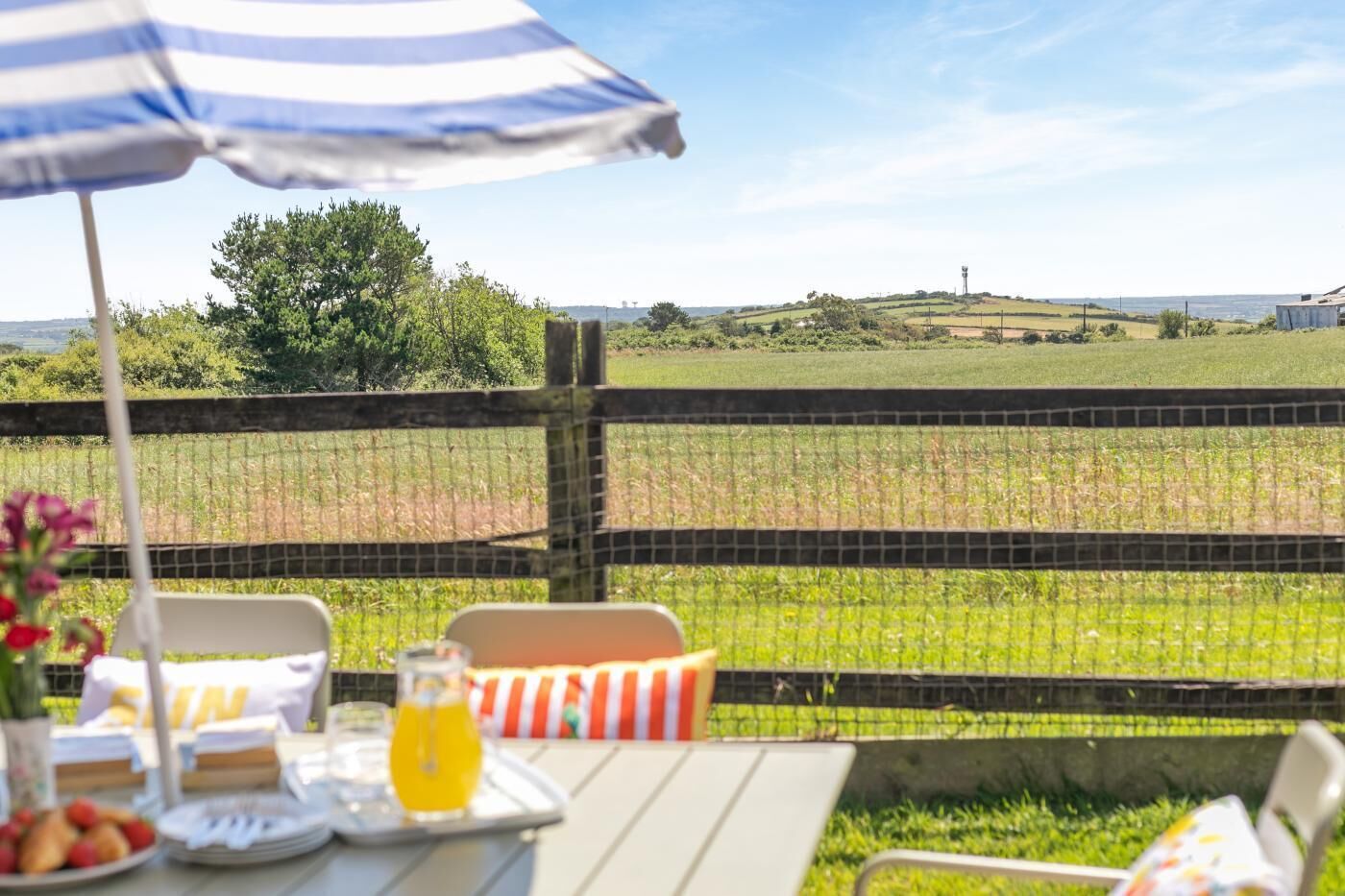 An outdoor dining area with a table set for two, a striped umbrella, and a view of a field and distant landscape.
