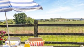 Uma área de refeições ao ar livre com uma mesa posta para dois, um guarda-sol listrado e vista para um campo e paisagem distante.
