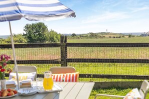Ein Essbereich im Freien mit einem Tisch für zwei Personen, einem gestreiften Sonnenschirm und Blick auf ein Feld und eine entfernte Landschaft.