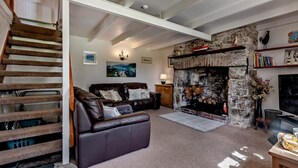 A living room with a stone fireplace, a brown leather sofa, a wooden staircase, and a bookshelf. - 2 Bed in Wadebridge (oc-jobes) (Chapel Amble)
