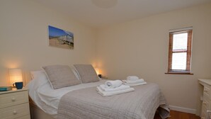 Ein Schlafzimmer mit einem Bett, Kissen, einem Nachttisch mit Lampe, einem Fenster mit Jalousien und einem Bild an der Wand.
