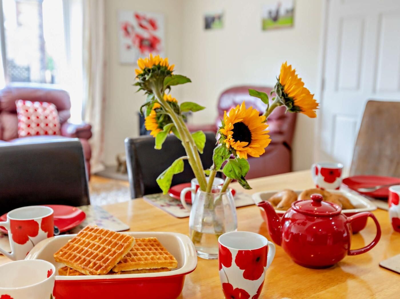 Een eettafel met rood en wit servies, een glazen vaas met zonnebloemen en een rode theepot.