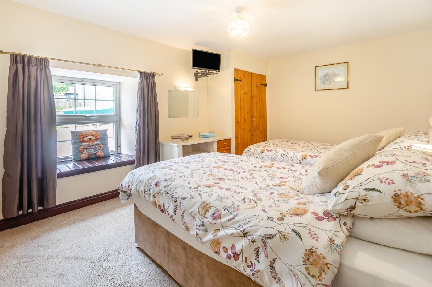 A bedroom with a bed, a window with curtains, a teddy bear, a bedside table, a mirror, a wooden wardrobe, and a wall-mounted TV.