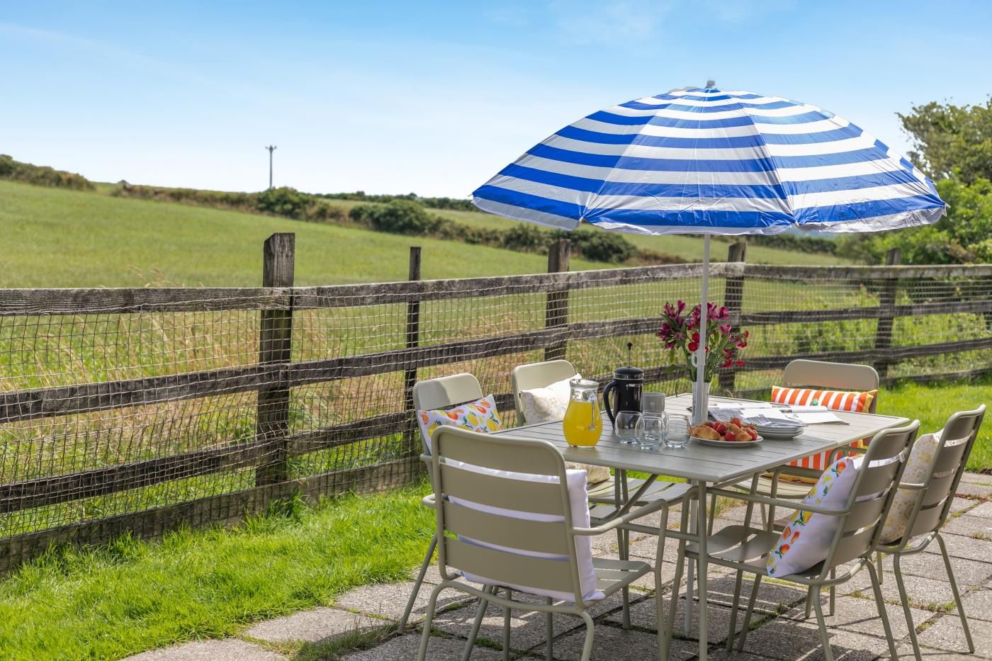 An outdoor dining area with a table, chairs, a striped umbrella, and a view of a grassy field.