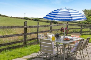 An outdoor dining area with a table, chairs, a striped umbrella, and a view of a grassy field. - 3 Bed in Falmouth (oc-consw) (Constantine)