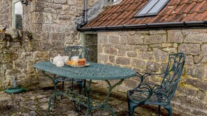 Un bâtiment en pierre avec un toit en tuiles, une table ronde en métal entourée de chaises et une petite fenêtre.