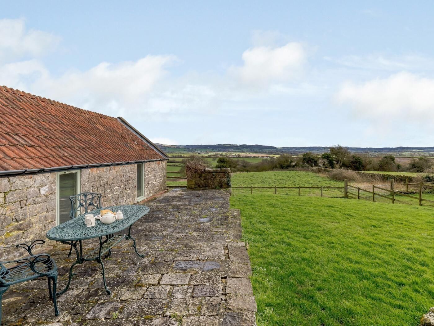 A stone building with a tiled roof, a stone patio with a table and chairs, and a grassy field with a fence in the background.