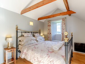 Ein Schlafzimmer mit Bett, Nachttisch, einem Fenster mit Vorhängen und freiliegenden Holzbalken.