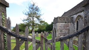 Une porte en bois rustique mène à un cimetière, avec un grand arbre en arrière-plan et un bâtiment en pierre doté de fenêtres en arc.
