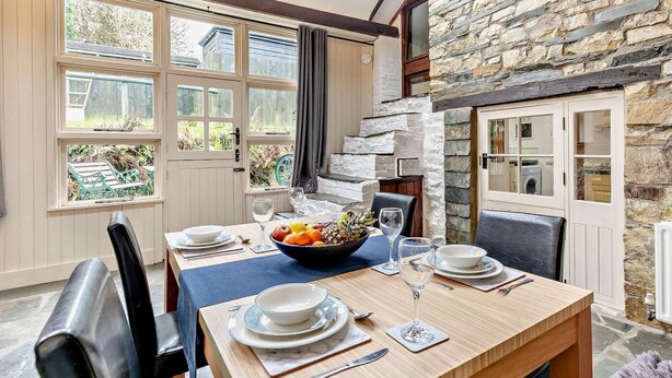 A dining area with a table set for four, a stone wall, and a view of the garden through large windows. - 1 Bed in Camelford (oc-hopar) (Lanteglos)