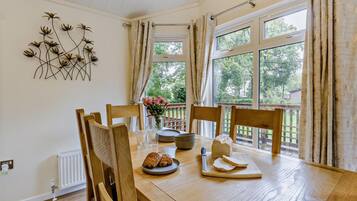 A dining area with a wooden table, chairs, a vase of flowers, and a view of greenery through large windows.