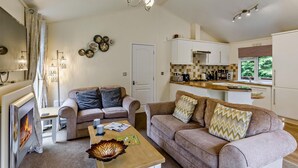 A living room with a fireplace, a sofa set, a coffee table, and a kitchen area with cabinets and a window. - 3 Bed in Okehampton (oc-06199) (Okehampton Hamlets)