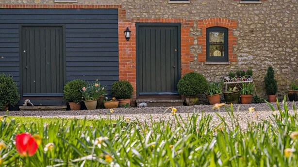 Een gebouw met twee donkergekleurde garage deuren, een stenen muur en een klein raam. Voor zijn er bloempotten en een metalen bank met bloemen.