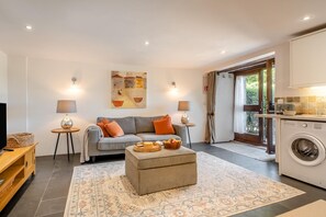 Ein modernes Wohnzimmer mit Sofa, Hocker und Teppich. In der Ecke steht eine Waschmaschine und es gibt einen Essbereich mit Tisch und Stühlen.