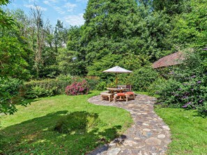 A stone pathway leading to a wooden outdoor seating area with a table and chairs, surrounded by lush greenery and flowering plants. - 4 Bed in Bideford (oc-hacot) (Hartland)