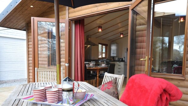 A wooden cabin with a dining area featuring a table with striped cups and a tray, a red towel, and a view into the kitchen. - 2 Bed in Redruth (oc-gillo) (Scorrier)