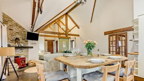 A dining area with a wooden table set for four, surrounded by wooden chairs with cushions. A stone wall and a fireplace are visible in the background. - 3 Bed in Camelford (oc-pendb) (Forrabury and Minster)