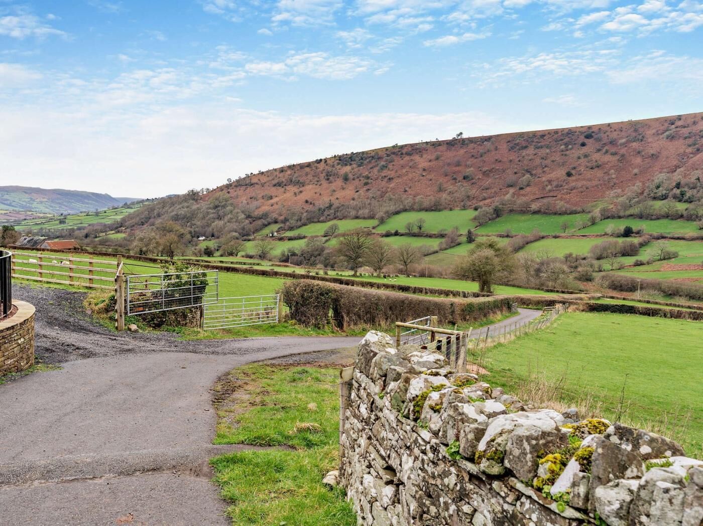 A rural landscape with a stone wall, a metal gate, and a winding road.