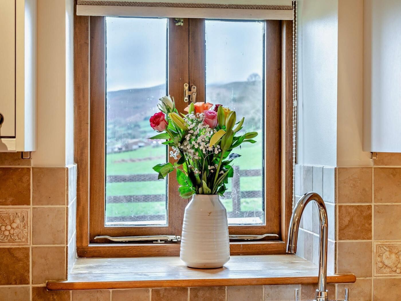 Une cuisine avec une fenêtre, un vase rempli de fleurs et une vue sur un paysage verdoyant.