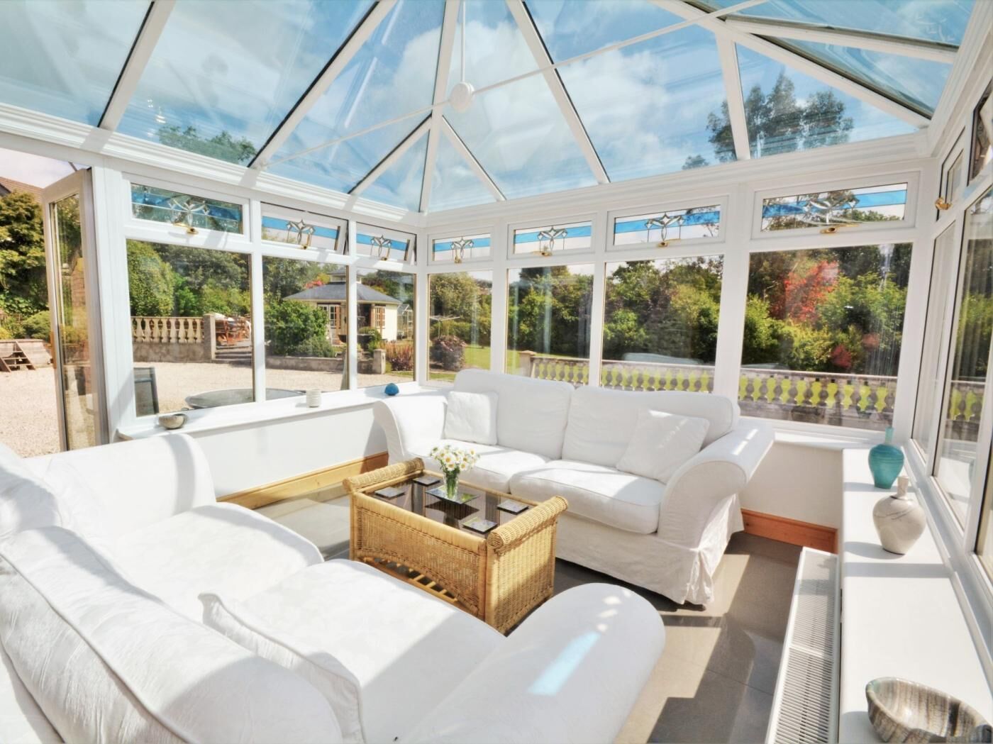 A conservatory with white furniture, a wicker coffee table, and a view of the garden.