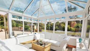 Uma estufa com móveis brancos, uma mesa de centro de vime e vista para o jardim.